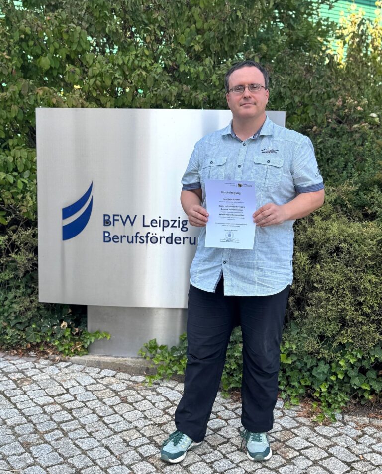 Junger Mann zeigt sein Abschlusszeugnis vor dem Eingangsschild des Berufsförderungswerkes Leipzig an der Georg-Schumann-Straße