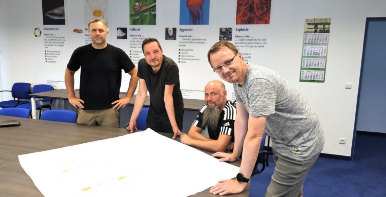 Vier Männer in einem Raum, mit einem Zeichenplan vor sich, unterhalten sich über neue Projekte. Sie sind alles Technische Produktdesigner.