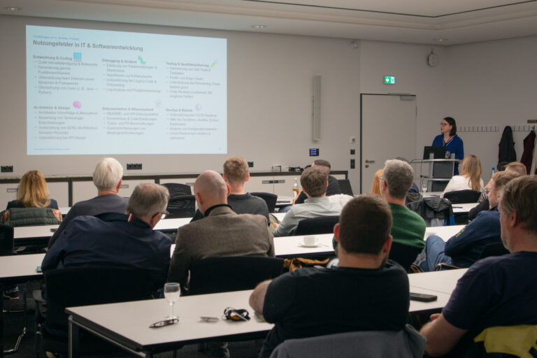 Blick in den Raum mit Referentin Sabine Maßmann, die vor den Gästen am Pult steht und zum Thema "Künstliche Intelligenz im Entwickleralltag" vor den Zuhörern referiert.