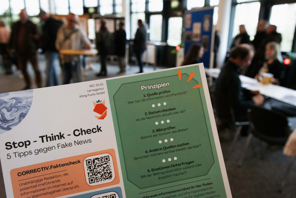 im Vordergrund: Kurzanleitung der Kaufleute im E-Commerce mit 5 Tipps gegen Fake News. Im Hintergrund sind unscharf die Besucher des Aktionstags zu sehen.