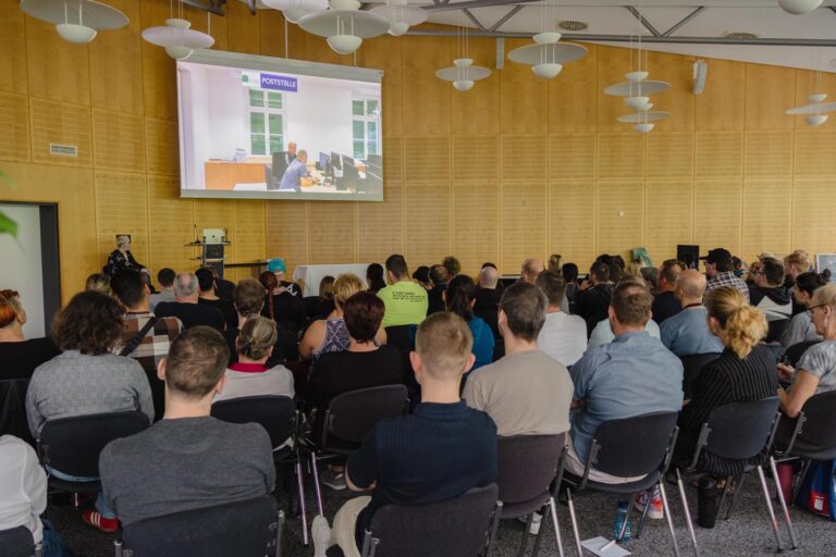 50 Personen sitzen auf Stühlen und schauen sich die Abschlusspräsentation des Lernunternehmens auf der großen Leinwand an
