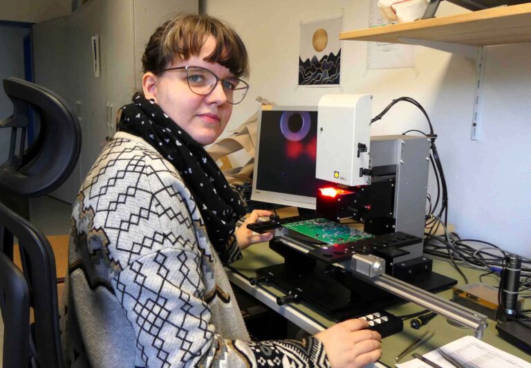 Eine weibliche Elektronikerin sitzt an einem Schreibtisch und prüft Platinen auf ihre Tauglichkeit.